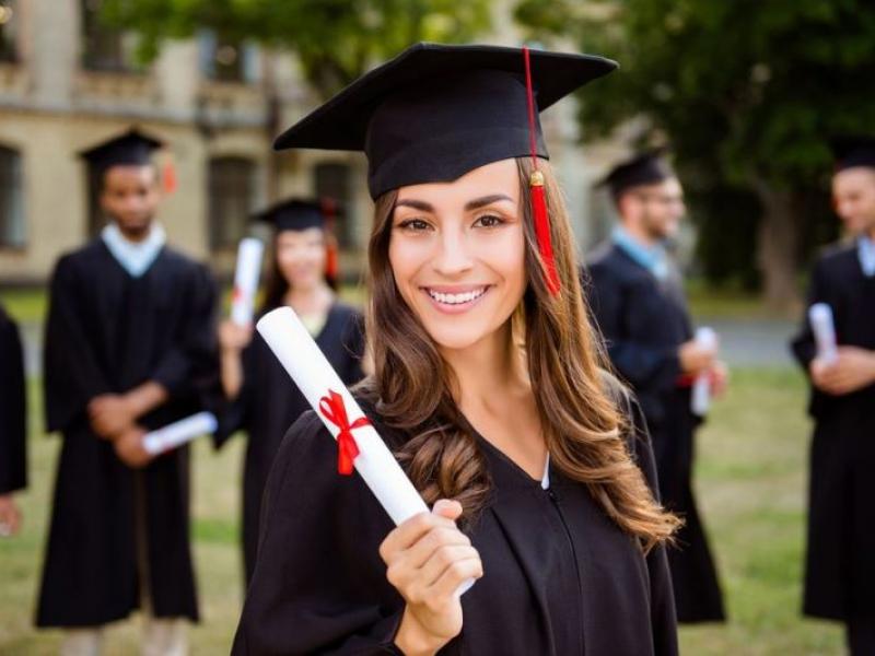 Zdjęcie przedstawia kobietę. Trzyma w ręku zwinięty w rulon biały dyplom. Rulon jest związany czerwoną wstążką. Kobieta ubrana jest w togę i biret absolwenta. W jej tle widać innych absolwentów.